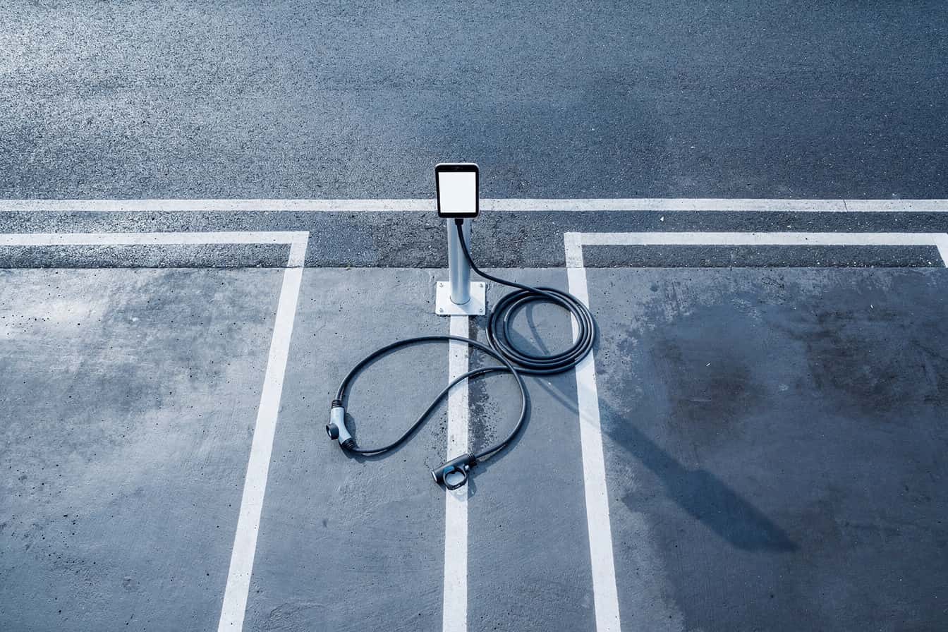A damaged public EV charging cable hangs from its connector in a Canterbury carpark at dusk, with an empty car park bay beside it.